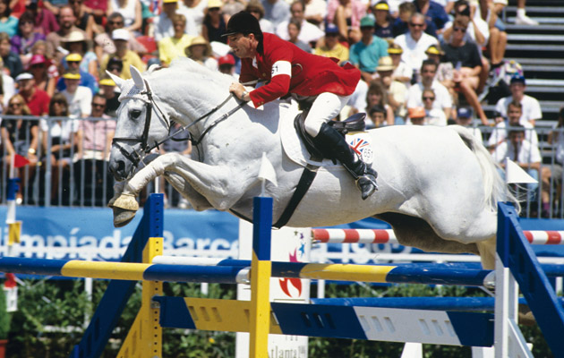 Une photo d'action d'archive du légendaire cavalier britannique John Whitaker montant son célèbre cheval gris, Milton, en plein saut au-dessus d'un grand obstacle de saut d'obstacles lors d'une compétition internationale de haut niveau. Milton est un grand gris au corps blanc uni, en pleine suspension au-dessus des barres bleues et jaunes. John Whitaker, portant une veste de concours rouge, des pantalons blancs et une bombe noire, est penché vers l'avant dans une posture de saut parfaite. Un drapeau de l'Union Jack est visible sur le tapis de selle et un dossard de compétition sur son bras. L'arrière-plan montre une grande foule de spectateurs floue dans un stade, avec des signes partiels pour 'Barcelona' et 'Atlanta', situant l'événement dans un contexte de compétition internationale de haut niveau.