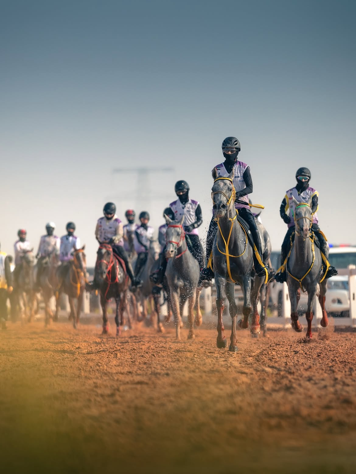 Peloton de cavaliers d'endurance équipés de casques, lunettes et cagoules de protection, galopant de front sur une piste de sable désertique. Au premier plan, un cheval gris mouillé par de l'eau mène la course.